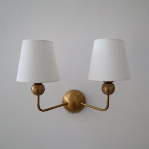 Peut inclure: Applique murale double avec une finition dorée. Elle comporte deux abat-jours blancs en forme de cône. Les abat-jours sont soutenus par des bras incurvés partant d'une sphère centrale dorée. Le luminaire est conçu pour l'éclairage intérieur.