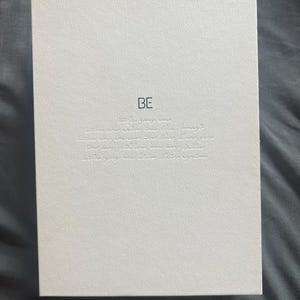 May include: A white rectangular box with the word "BE" printed in black at the top. Below the word "BE" is a series of embossed text. The box has a textured surface and is resting on a gray surface.