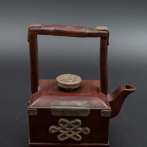 May include: A reddish-brown wooden teapot with a handle and spout. The teapot has decorative metal accents on the corners and a circular lid. An ornate metal design is on the front.
