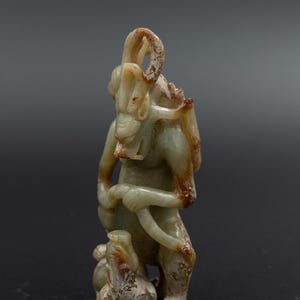 以下が含まれることがあります： 角のある頭と足元に小さな生き物がいる立像を描いた、彫刻されたヒスイの置物。 フィギュアは、暗い背景に対して、茶色のアクセントが付いた明るい緑色をしています。