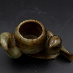 May include: A carved, olive-green stone vessel with a circular opening and stylized bird-like form. The sculpture has a smooth, polished surface with subtle variations in color, and is set against a dark background.