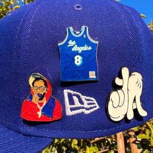 Può includere: Un berretto da baseball blu reale adornato con spille smaltate. Le spille includono una maglia dei Los Angeles Lakers, un logo New Era, una figura di cartone animato e un gesto della mano in bianco e nero.