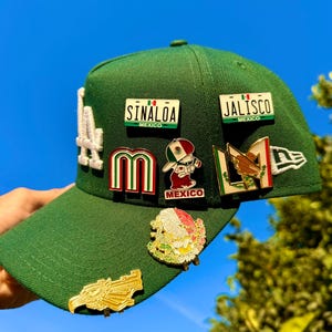 Può includere: Cappellino da baseball verde con diverse spille smaltate. Le spille includono i testi "SINALOA MEXICO" e "JALISCO MEXICO", insieme ad altri disegni a tema messicano. Il cappellino ha un logo "LA" bianco sul lato.