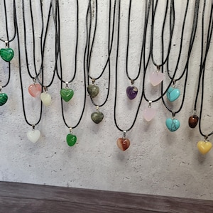 May include: A collection of heart-shaped gemstone pendants on black cords. The pendants vary in color, including green, pink, purple, and blue. Each necklace has a silver-colored clasp. The background is a light gray wall.