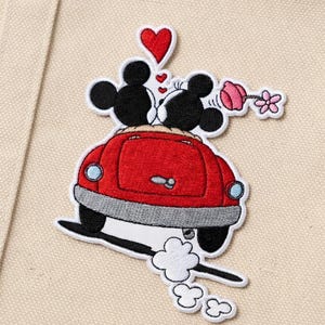 May include: Embroidered patch featuring Mickey and Minnie Mouse kissing in a red car, with a heart above them. The patch includes a flower and exhaust smoke, set against a beige background.