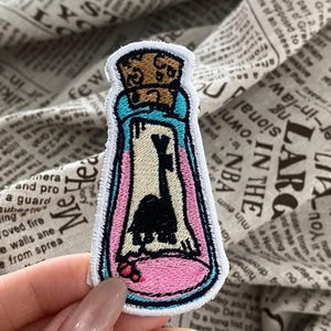 Puede incluir: Parche bordado que representa una botella con una silueta negra en su interior, sobre un fondo rosa y azul. El parche tiene un tapón de corcho y un borde blanco. Una pequeña flor roja está en la parte inferior.
