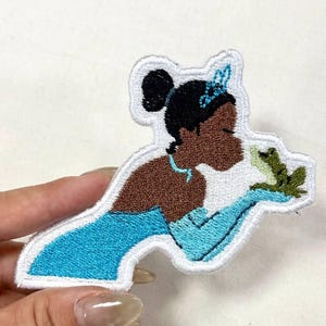 Op de afbeelding: Geborduurde patch met een prinses in een blauwe jurk die een kikker vasthoudt. De patch heeft een witte rand en is gemaakt met blauw, bruin, zwart en groen garen. De prinses draagt een tiara.