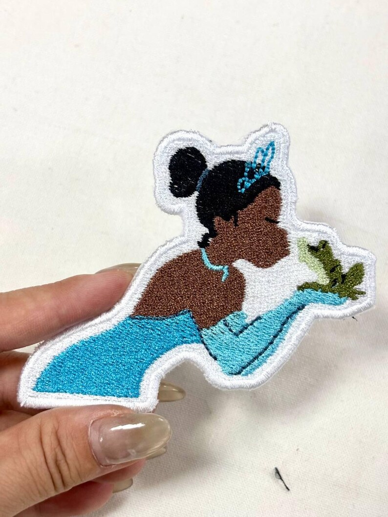 Op de afbeelding: Geborduurde patch met een prinses in een blauwe jurk die een kikker vasthoudt. De patch heeft een witte rand en is gemaakt met blauw, bruin, zwart en groen garen. De prinses draagt een tiara.