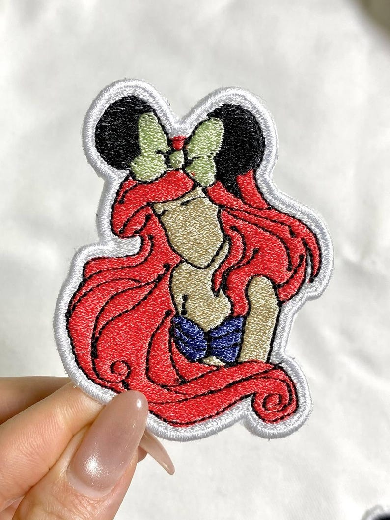 May include: Embroidered patch featuring a cartoon character with red hair, a blue top, and a green bow. The patch has a white border and is likely for clothing or accessories.