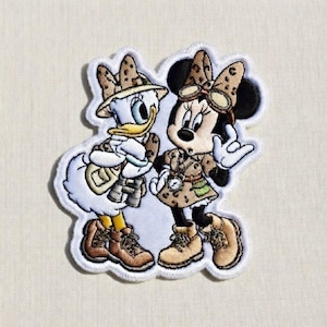Peut inclure: Écusson brodé représentant Daisy Duck et Minnie Mouse en tenues de safari. Daisy porte un chapeau de safari, des jumelles et une sacoche. Minnie a des lunettes et une robe à imprimé léopard. Les deux personnages portent des bottes de randonnée. Le patch a une bordure blanche.