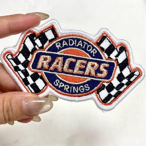 May include: Embroidered patch featuring the text "RADIATOR RACERS SPRINGS" in a circular design with two checkered flags. The patch has a white border, with orange, blue, and black accents.