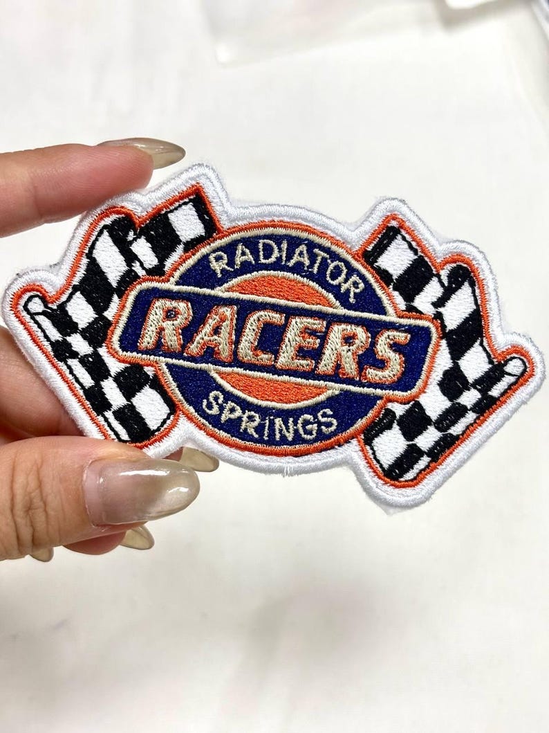 May include: Embroidered patch featuring the text "RADIATOR RACERS SPRINGS" in a circular design with two checkered flags. The patch has a white border, with orange, blue, and black accents.
