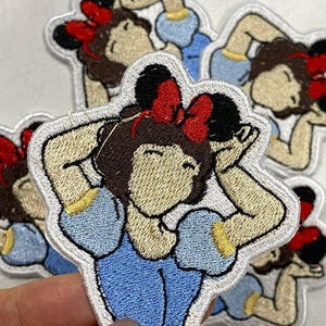May include: Embroidered patch featuring a cartoon character with brown hair, a red bow, and a blue dress. The patch has a white border and is likely for clothing or accessories.