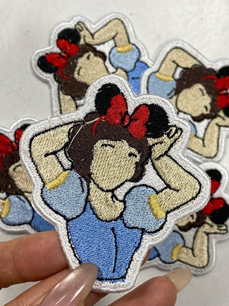 May include: Embroidered patch featuring a cartoon character with brown hair, a red bow, and a blue dress. The patch has a white border and is likely for clothing or accessories.
