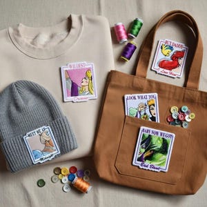 May include: A tan sweatshirt and a gray beanie with embroidered patches featuring cartoon characters. A brown tote bag with patches and buttons is also present. Spools of thread in various colors are scattered around the items.