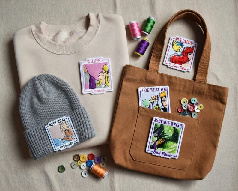 May include: A tan sweatshirt and a gray beanie with embroidered patches featuring cartoon characters. A brown tote bag with patches and buttons is also present. Spools of thread in various colors are scattered around the items.