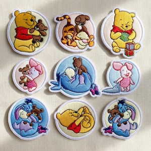 Könnte beinhalten: Neun gestickte Aufnäher mit Winnie Puuh und seinen Freunden. Die Aufnäher zeigen Charaktere wie Puuh, Ferkel, Tiger und Eeyore, jeweils in einem kreisförmigen Design mit weißem Rand. Die Aufnäher sind farbenfroh, mit gelben, roten, rosa und blauen Farbtönen.