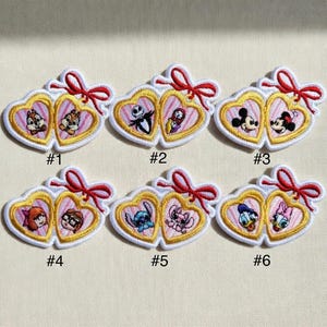 Op de afbeelding: Geborduurde patches met paren Disney-personages in hartvormige frames. Elke patch heeft een gouden rand, witte omtrek en een rode strik. De patches zijn genummerd van &eacute;&eacute;n tot zes en tonen personages als Chip en Dale.