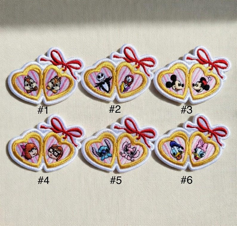 Op de afbeelding: Geborduurde patches met paren Disney-personages in hartvormige frames. Elke patch heeft een gouden rand, witte omtrek en een rode strik. De patches zijn genummerd van &eacute;&eacute;n tot zes en tonen personages als Chip en Dale.