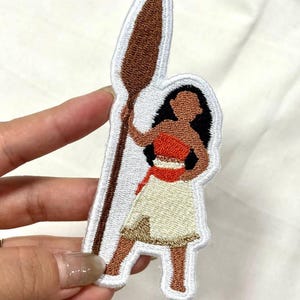 Op de afbeelding: Geborduurde patch met een Polynesische vrouw die een speer vasthoudt. De patch heeft een witte rand en toont de vrouw in een rode top, cr&egrave;mekleurige rok en bruine huid. De speer is bruin en de vrouw heeft zwart haar.