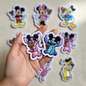 May include: Embroidered patches featuring various cartoon characters, including Mickey Mouse, Minnie Mouse, Donald Duck, Stitch, and Angel. The patches have white borders and are in various colors. Some patches are held in a hand.