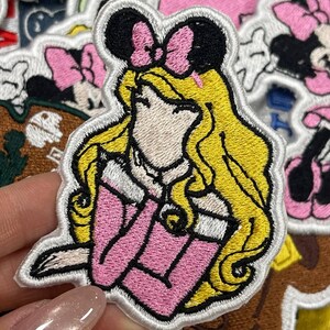 May include: Embroidered patch featuring a princess with blonde hair, wearing a pink dress and a black bow. The patch has a white border and is surrounded by other patches with cartoon characters. Ideal for decorating clothing or accessories.