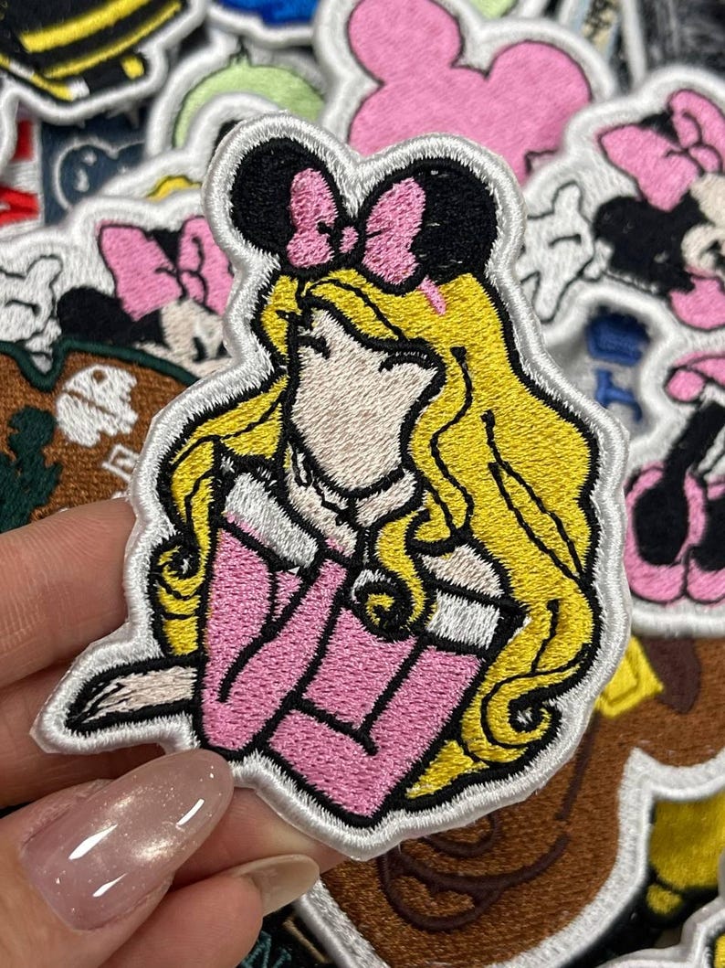 May include: Embroidered patch featuring a princess with blonde hair, wearing a pink dress and a black bow. The patch has a white border and is surrounded by other patches with cartoon characters. Ideal for decorating clothing or accessories.