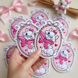 May include: Embroidered patches featuring a white cat with a pink bow, set against a pink oval frame. The patches have a cartoon design with a pink background and a small red heart. A small figurine and pink bows are also visible.