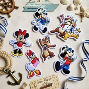 Pode incluir: Patches bordados com personagens da Disney, Mickey e Minnie Mouse, Pato Donald e Tico e Teco, em trajes náuticos. Os patches brancos têm detalhes coloridos e são dispostos com decorações náuticas.