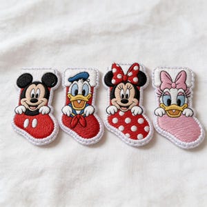 May include: Four embroidered iron-on patches shaped like Christmas stockings, each featuring a different Disney character: Mickey Mouse in a red stocking, Donald Duck in a red stocking, Minnie Mouse in a red polka-dot stocking, and Daisy Duck in a pink stocking.