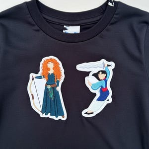 Op de afbeelding: Zwart sweatshirt met ronde hals en twee geborduurde patches. E&eacute;n patch toont een vrouw met rood krullend haar die een pijl en boog vasthoudt. De andere patch toont een vrouw die een zwaard vasthoudt.