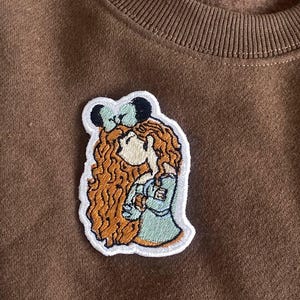 May include: Embroidered patch featuring a cartoon illustration of a girl with long, curly auburn hair and a bow. The patch has a white border and is sewn onto a brown sweatshirt. The girl is wearing a light blue dress.