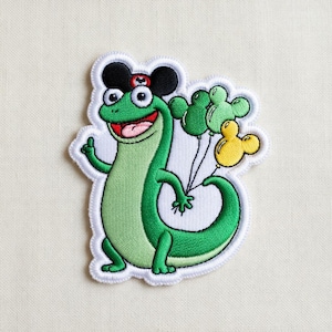 May include: Embroidered patch featuring a cartoon lizard with a cheerful expression. The lizard is green with a red mouth and is wearing a black and red hat. It holds green and yellow Mickey Mouse-shaped balloons. The patch has a white border.