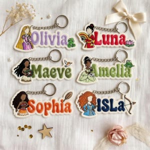 May include: Six embroidered keychains featuring cartoon characters and names: Olivia in purple, Luna in red, Maeve in green, Amelia in light green, Sophia in orange, and Isla in blue. Each keychain includes a metal ring and character-themed icons.