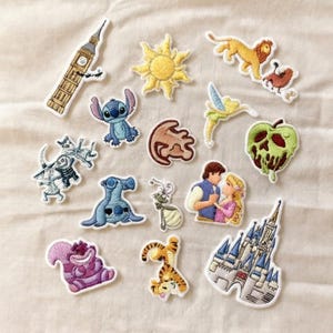 Può includere: Una collezione di toppe ricamate con vari personaggi dei cartoni animati e simboli iconici. Le toppe includono Stitch blu, un sole giallo, una mela verde, un leone, un castello e altri disegni. Le toppe hanno bordi bianchi e sembrano fatti a mano.