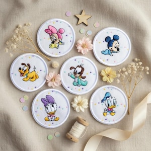 Puede incluir: Parches circulares bordados con personajes de dibujos animados, incluyendo Mickey Mouse, Minnie Mouse, Donald Duck, Daisy Duck, Pluto y Goofy. Cada parche es blanco con un diseño colorido del personaje y un borde punteado. También hay flores decorativas y una estrella de madera.