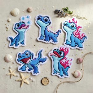 May include: Set of five embroidered patches featuring blue lizard-like creatures with varying expressions. Some have flames, snowflakes, or their tongues out. Patches are outlined in white, with a variety of seashells and a button surrounding them.