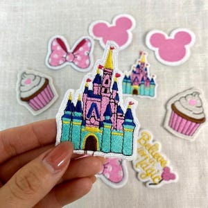 May include: A collection of embroidered patches featuring a pink and blue castle, pink mouse ears, cupcakes, and a pink bow with white polka dots. One patch reads "Believe in Magic".