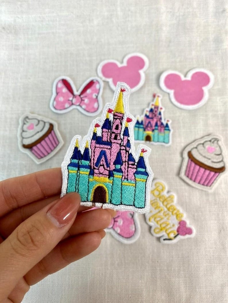 May include: A collection of embroidered patches featuring a pink and blue castle, pink mouse ears, cupcakes, and a pink bow with white polka dots. One patch reads "Believe in Magic".