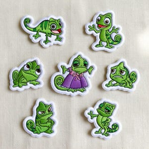 Op de afbeelding: Zeven geborduurde patches met een groene kameleon in verschillende poses. Eén kameleon draagt een paarse jurk. Elke patch heeft een witte rand en staat op een neutrale achtergrond.