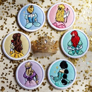 May include: Embroidered patches featuring various princess designs in a circular shape. Each patch showcases a different princess with detailed stitching and vibrant colors. A gold-colored jeweled crown is also present.