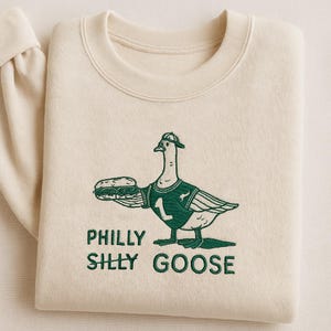 Könnte beinhalten: Cremefarbenes Sweatshirt mit grünem Stickdesign. Das Design zeigt eine Gans mit einem Football-Trikot und einem Sandwich, darunter die Worte "PHILLY SILLY GOOSE".