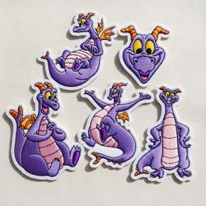 May include: A collection of six embroidered patches featuring a purple dragon character with orange horns and wings. The dragon has a pink belly and is in various poses, including sitting, smiling, and with arms outstretched. The patches have a white border.