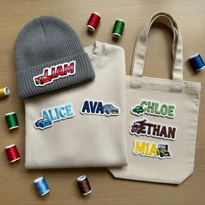 Puede incluir: Un gorro gris con el nombre "LIAM" y un parche de coche de carreras rojo. Una sudadera y un bolso tote beige presentan parches con nombres con gr&aacute;ficos de coches. Bobinas de hilo de varios colores est&aacute;n esparcidas alrededor de los art&iacute;culos.