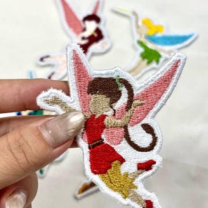 Op de afbeelding: Geborduurde fee&euml;npatches met roze vleugels, een rode jurk en bruin haar. De patches hebben een witte rand en staan op een witte achtergrond. Andere fee&euml;npatches zijn zichtbaar op de achtergrond.