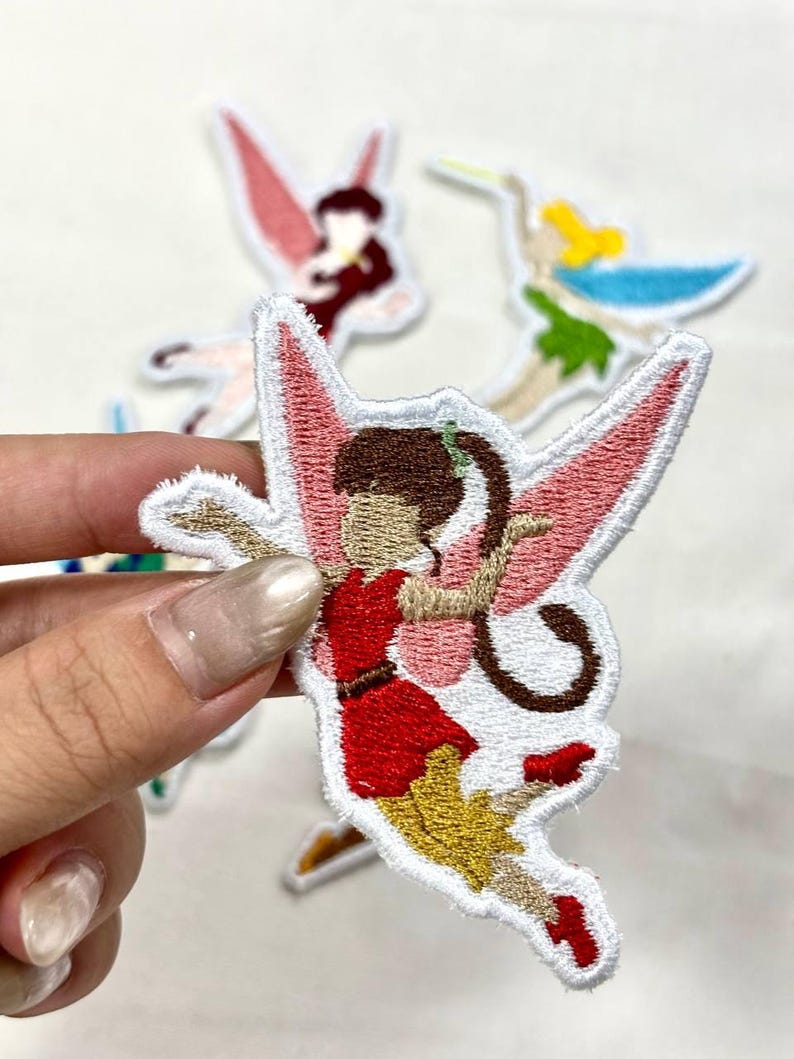 Op de afbeelding: Geborduurde fee&euml;npatches met roze vleugels, een rode jurk en bruin haar. De patches hebben een witte rand en staan op een witte achtergrond. Andere fee&euml;npatches zijn zichtbaar op de achtergrond.