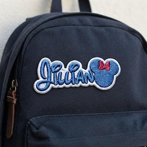 May include: A navy blue backpack featuring an embroidered patch with the name Lillian in blue script lettering. The patch includes a blue mouse-ear silhouette with a small red bow, all outlined in white thread against the dark fabric surface.