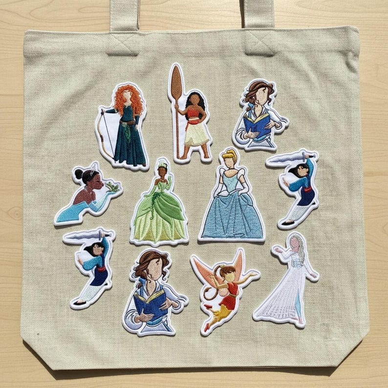 Op de afbeelding: Beige draagtas met geborduurde patches van verschillende geanimeerde vrouwelijke personages. De patches tonen personages in kleurrijke jurken en outfits, sommige met voorwerpen als boeken en zwaarden. De tas heeft twee handvatten.