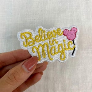 May include: Embroidered patch with the words "Believe in Magic" in yellow script. A pink Mickey Mouse head balloon is on the right. The patch has a white border and is on a white fabric background.