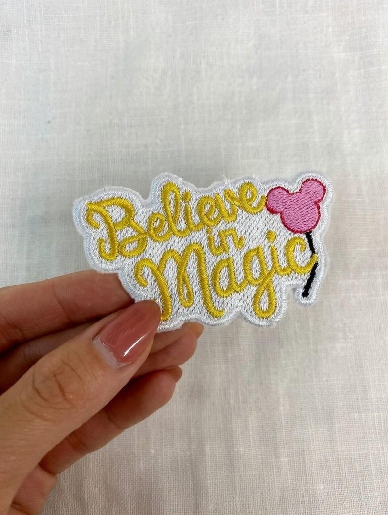 May include: Embroidered patch with the words "Believe in Magic" in yellow script. A pink Mickey Mouse head balloon is on the right. The patch has a white border and is on a white fabric background.